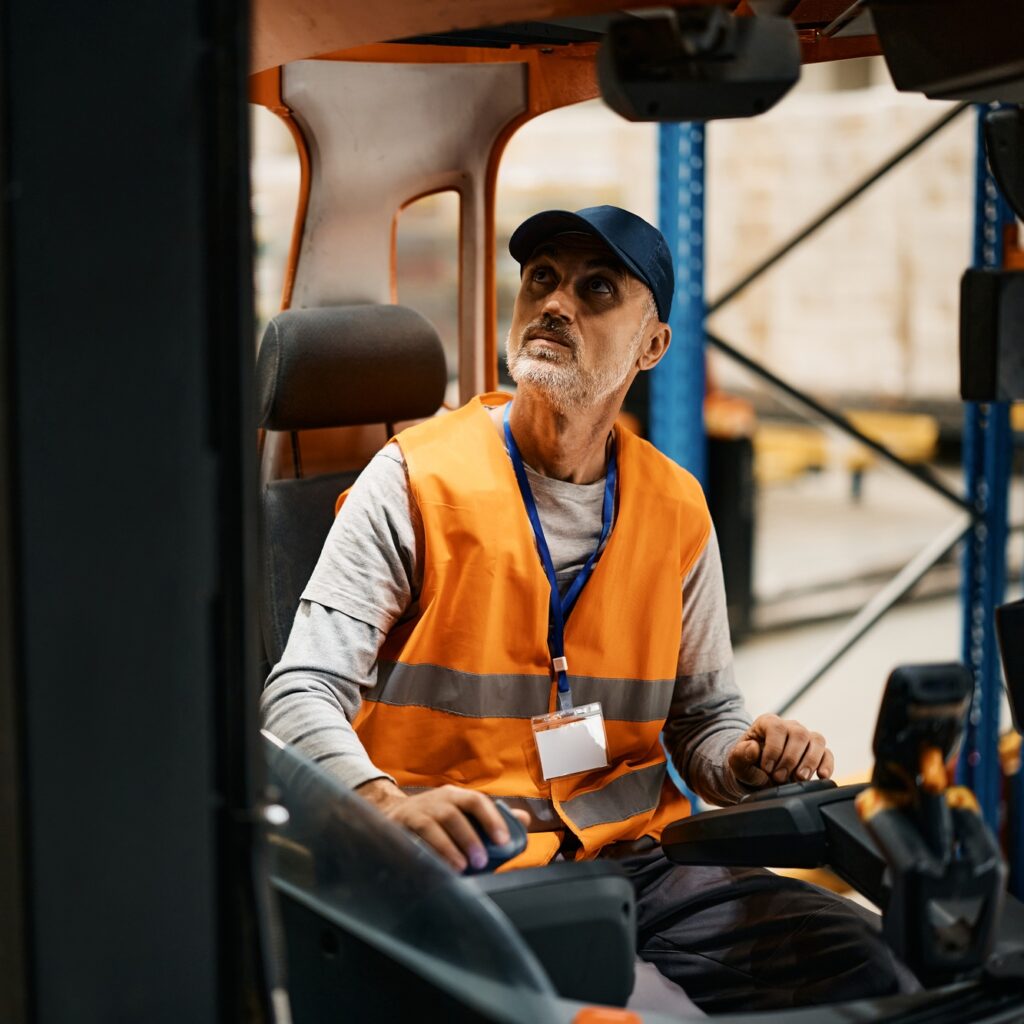 Mature warehouse worker moving pallets with forklift in industrial building.